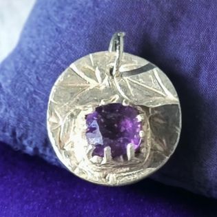 A round sterling silver pendant with a textured, leaf-like pattern and a central square-cut purple amethyst gemstone, resting on a textured purple fabric background.