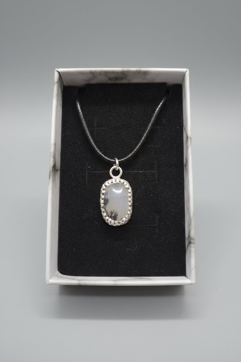 A silver pendant necklace with a semi-translucent oval stone, displayed inside an open white marble-patterned gift box with a black velvet insert.
