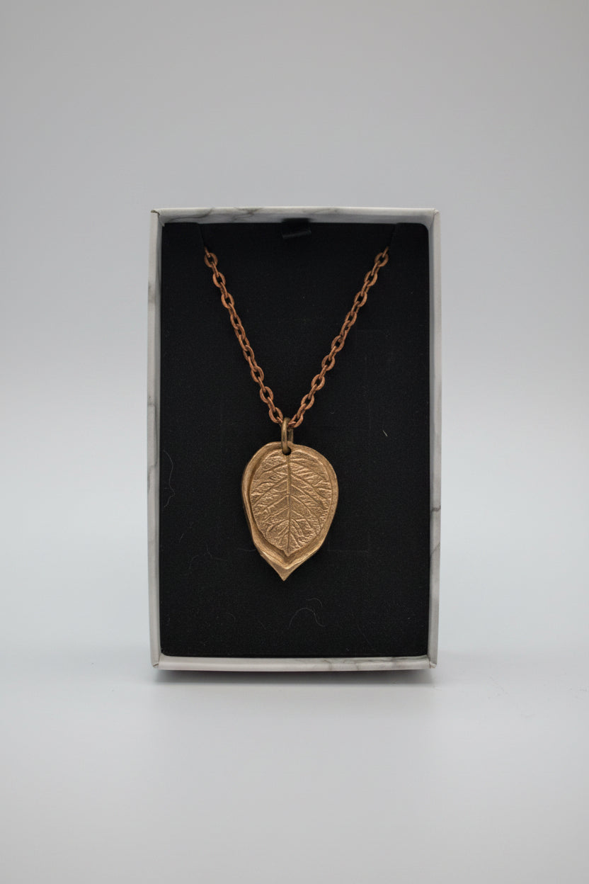 A copper-toned leaf pendant necklace on a matching chain, displayed inside an open white marble-patterned gift box with a black velvet lining.