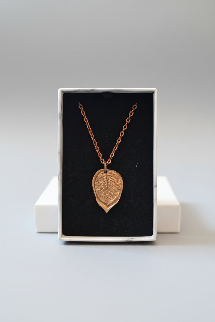 A copper-colored leaf pendant necklace displayed on a black velvet insert inside a white box.