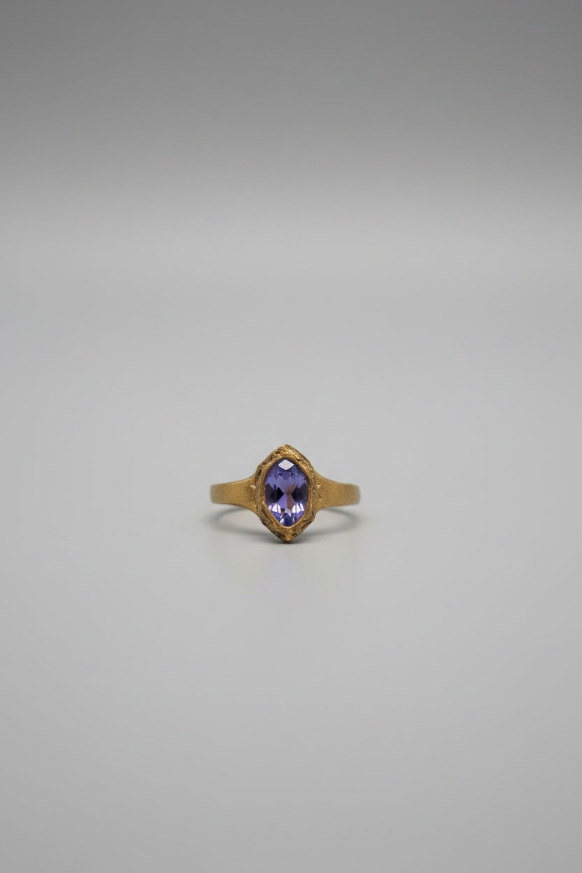 A gold ring with a oval-cut purple gemstone displayed on a black-lined 9-grid jewelry tray.