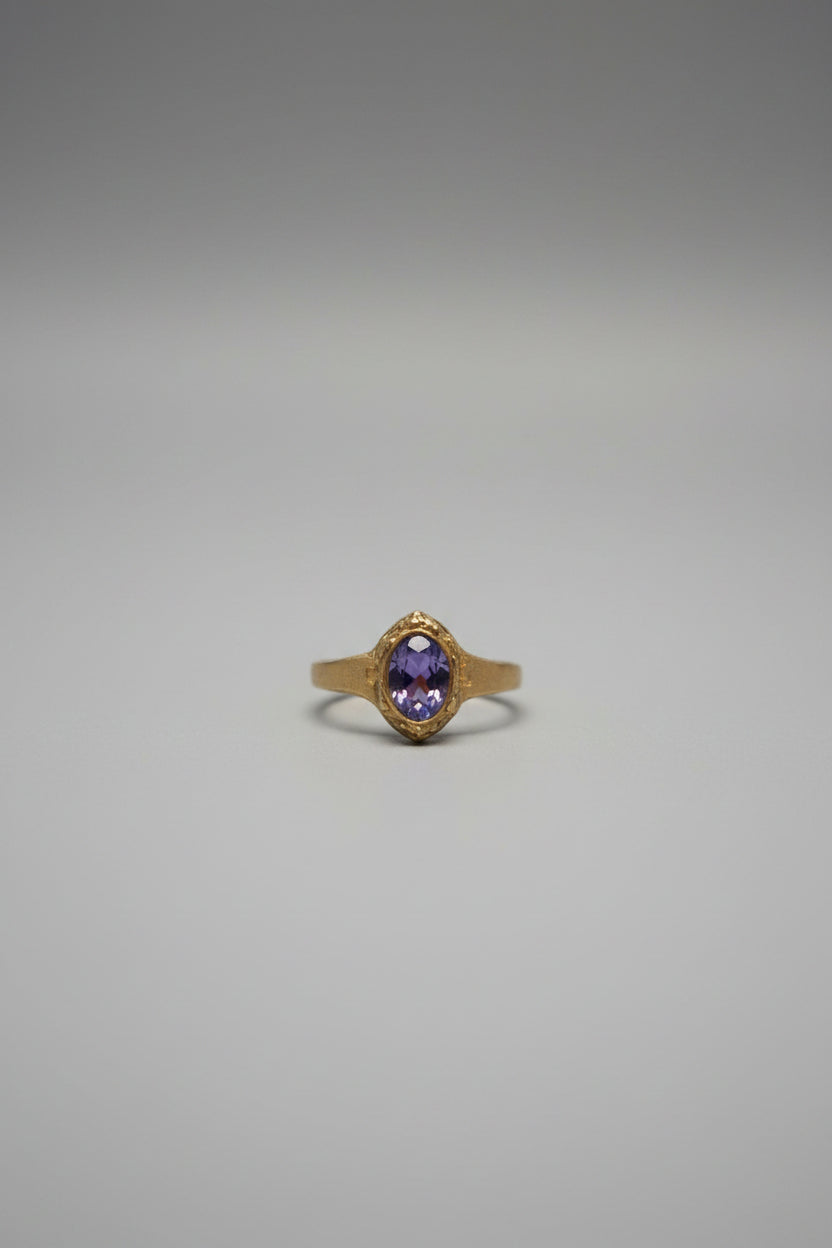 A gold ring with a purple oval gemstone, displayed on a black cushion inside a wooden jewelry organizer.