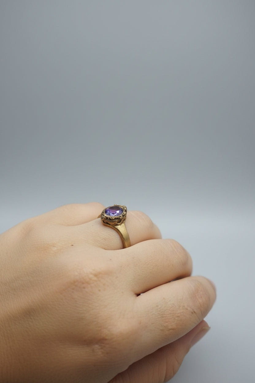 A gold ring with a purple gemstone worn on a person's finger, photographed in a real indoor setting with a jewelry tray blurred in the background.