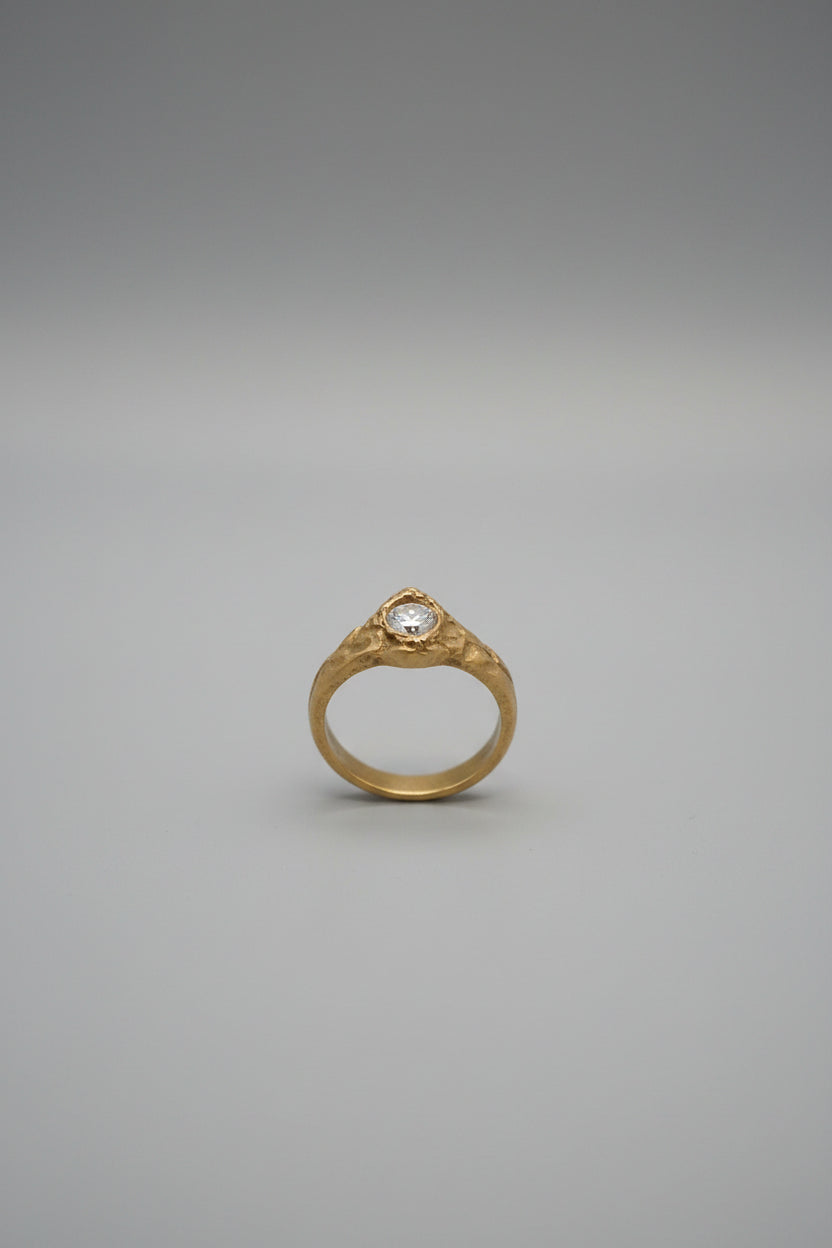 A bronze ring with a clear round gemstone set in an ornate band, displayed on a black ring stand within a light wood jewelry organizer.
