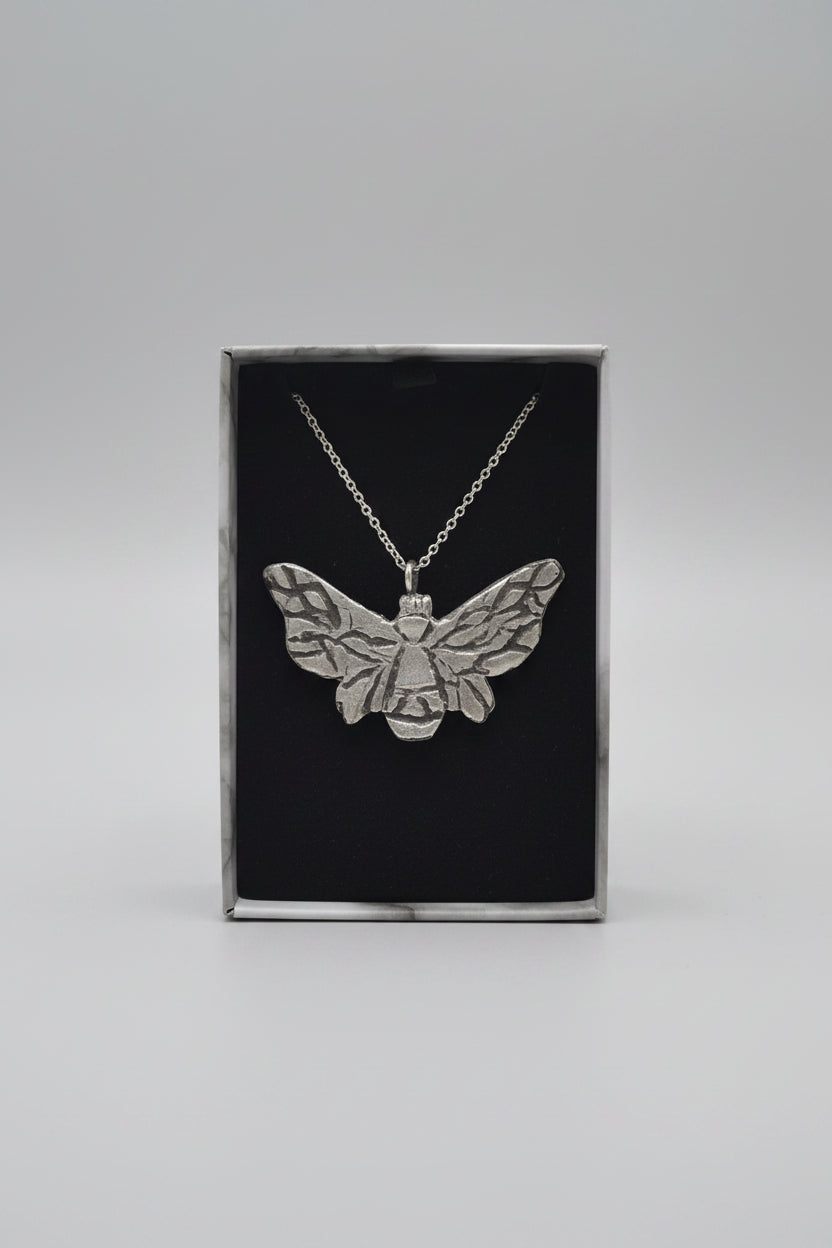 A silver bumble bee pendant necklace displayed inside a black presentation box, held by a hand with a laptop blurred in the background.