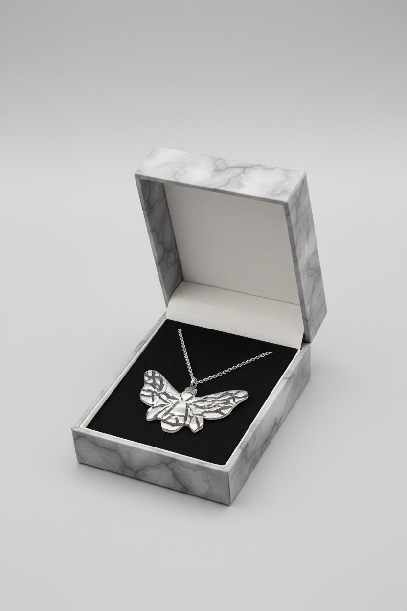 A bumble bee pendant necklace with a textured, hammered silver finish hangs from a delicate silver chain inside an open black presentation box, held by a hand.