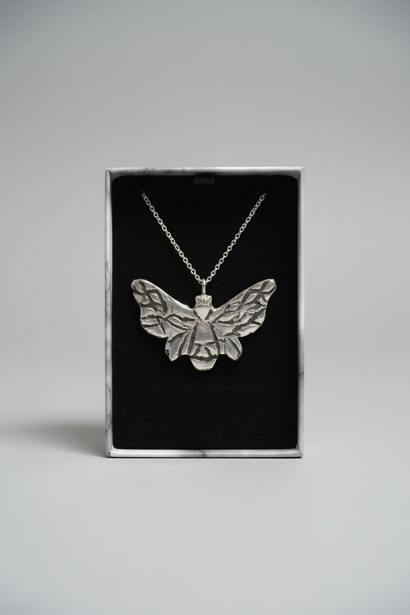 A silver-toned bee pendant necklace displayed inside an open black gift box, held by a hand with a blurred laptop in the background.