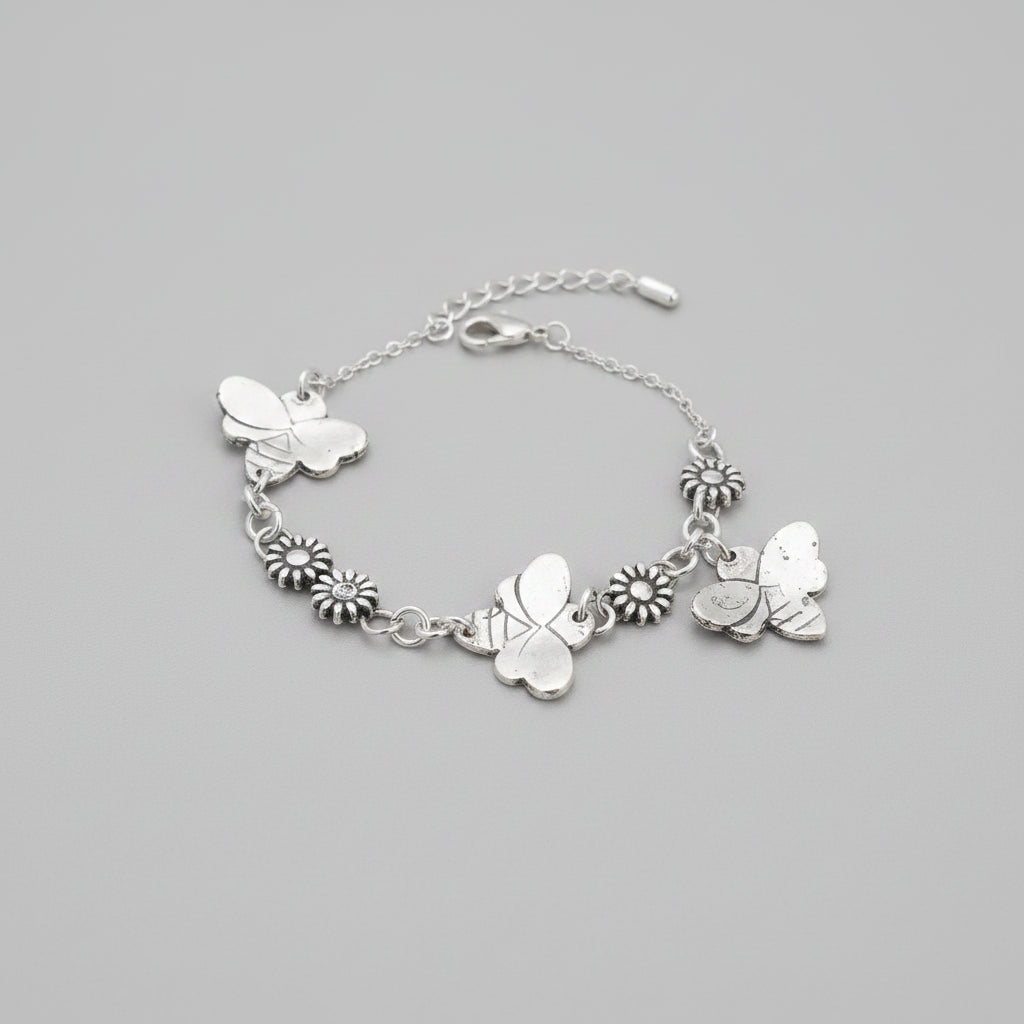 A silver-toned chain bracelet with butterfly and flower charms, displayed on a textured gray fabric surface with a wooden floor in the background.