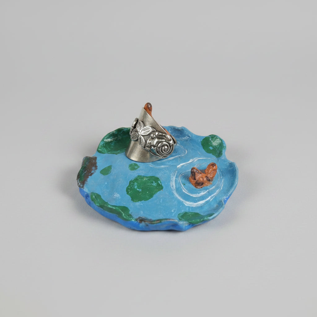 A miniature decorative figurine of a small boat on blue water with green patches, placed on a patterned cushion in a home setting.