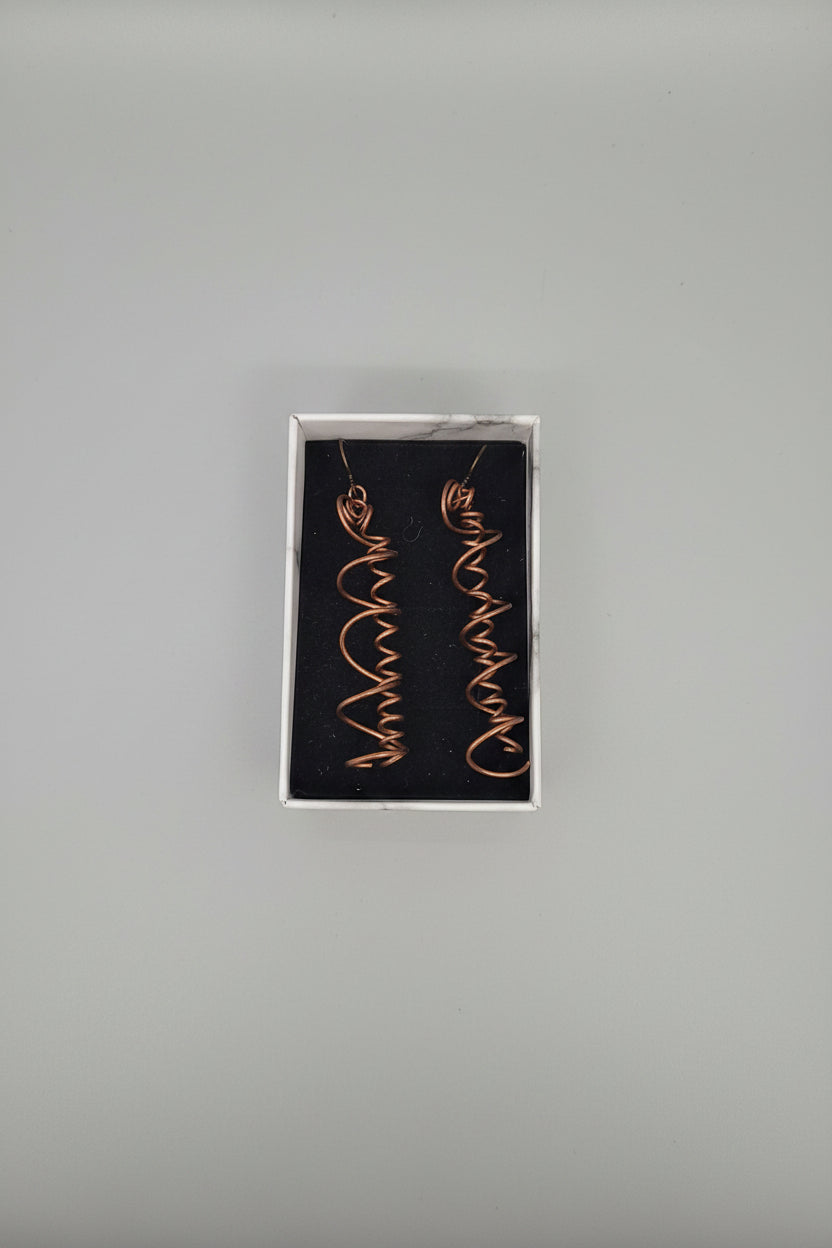 A pair of copper-colored spiral drop earrings displayed inside a small white jewelry box with a black velvet insert, set on a gray textured fabric surface.