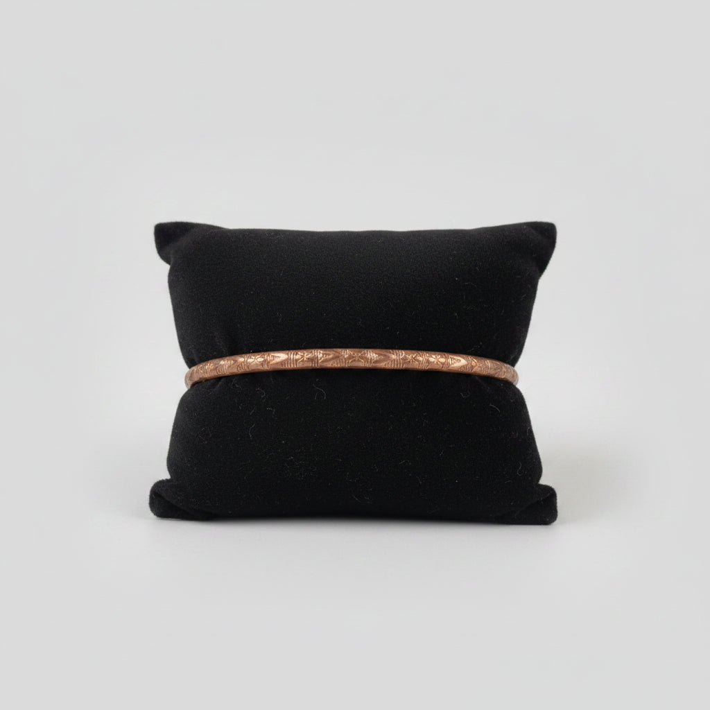 A thin copper bangle bracelet with a textured pattern, displayed on a black velvet jewelry pillow in a home setting.
