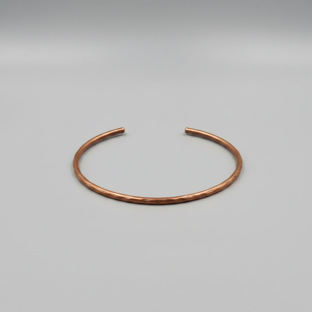 A single open cuff bracelet made of polished copper-colored metal, displayed on a textured gray fabric background.