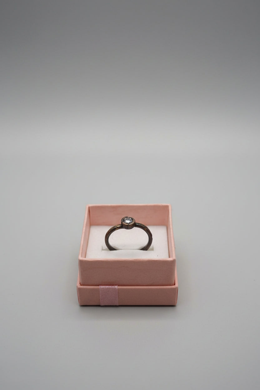 A vintage-style ring with a dark oxidized metal band and a round clear gemstone, presented inside a small pinkish-brown jewelry box on a desk with a keyboard in the background.