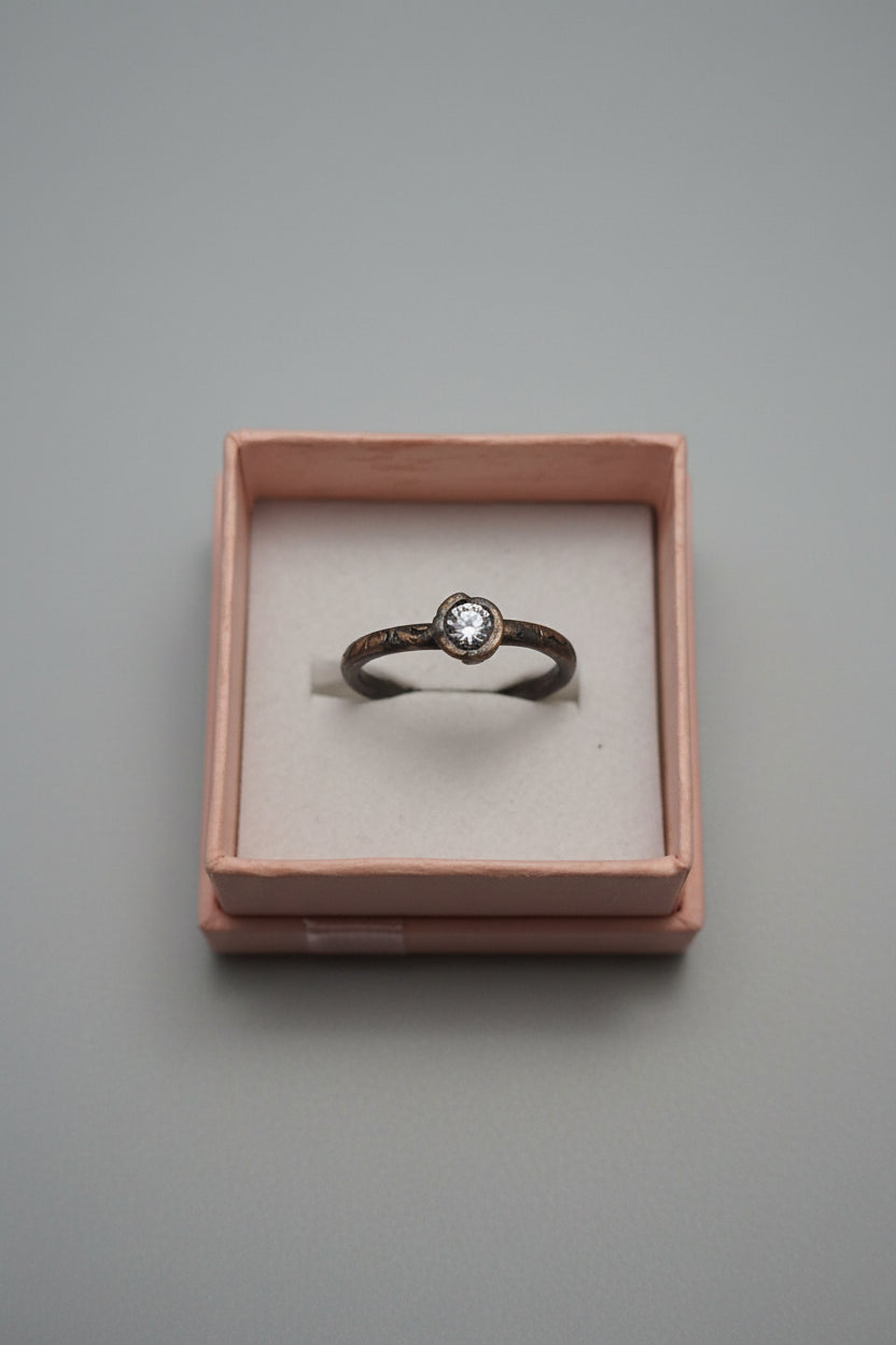 A vintage-style ring with a dark oxidized band and a small clear gemstone, displayed inside a pinkish-brown jewelry box with a keyboard in the blurred background.