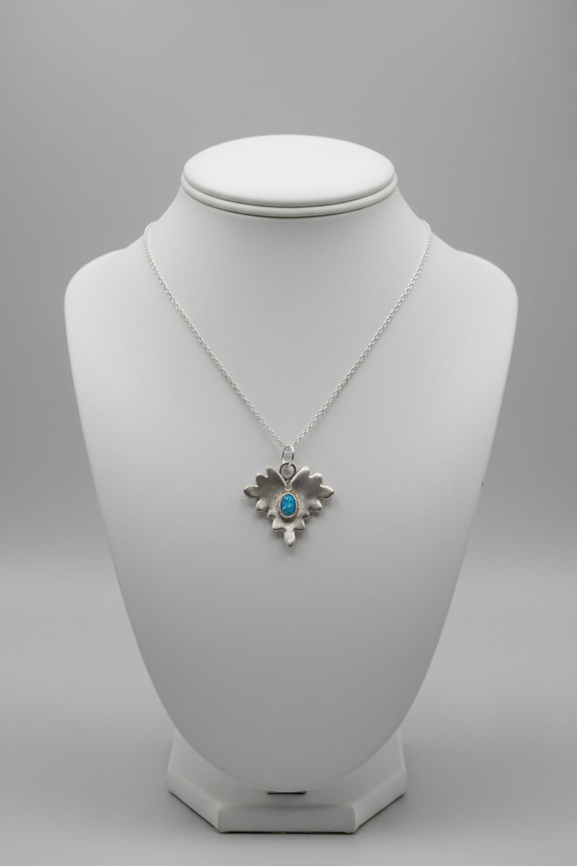 A silver-toned pendant necklace with a textured, organic leaf or flame-like shape featuring a small oval dark blue stone in the center, displayed on a burlap-covered mannequin bust.