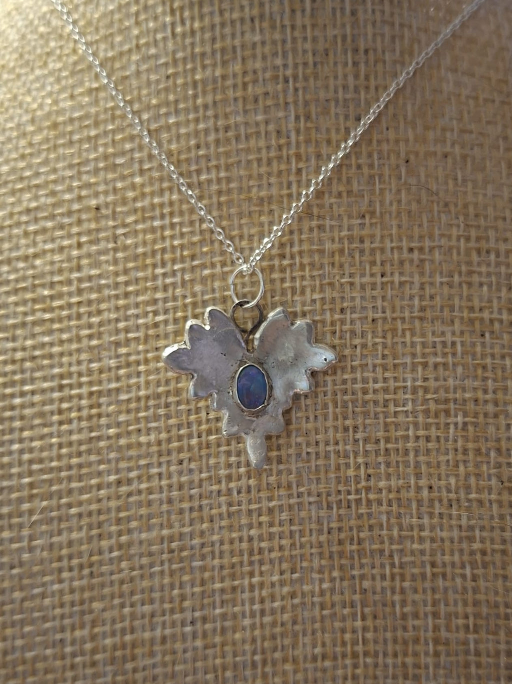 A silver-toned pendant necklace with a textured, leaf-like or flame-like silhouette and a small oval blue stone in the center, displayed on a burlap-covered jewelry bust.