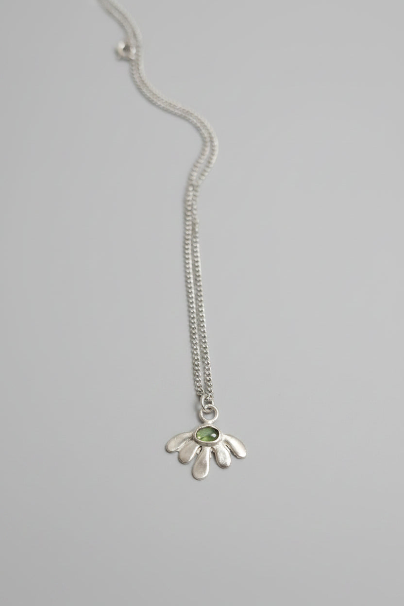 A sterling silver flower pendant necklace with a small green gemstone in the center, displayed on a textured dark brown leather surface.