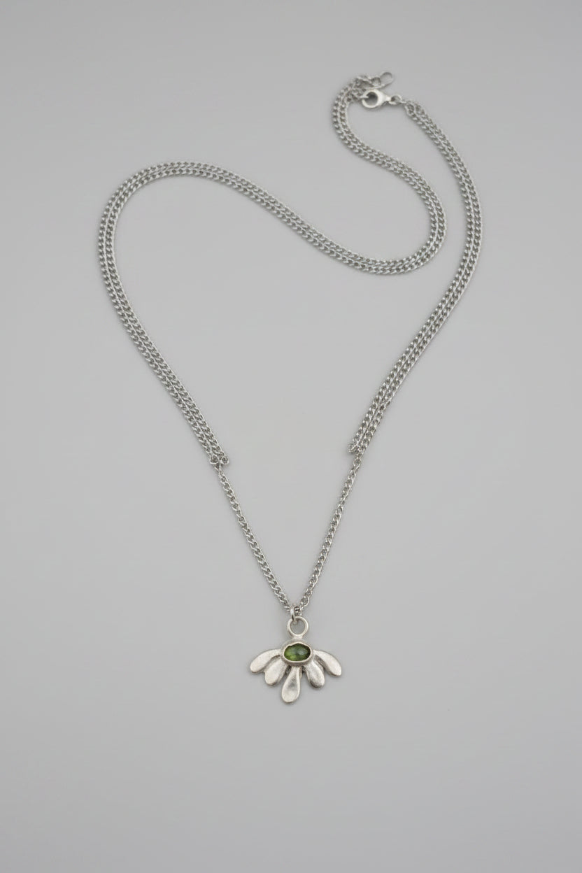 A silver-toned flower pendant necklace with a small green gemstone in the center, displayed on a textured dark brown surface.