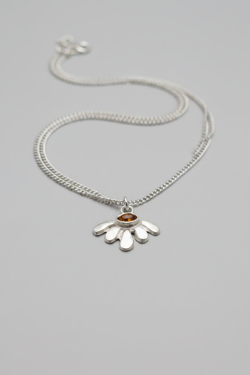 A silver-toned pendant necklace featuring a stylized flower with a small amber-orange center, resting on a textured dark brown surface.