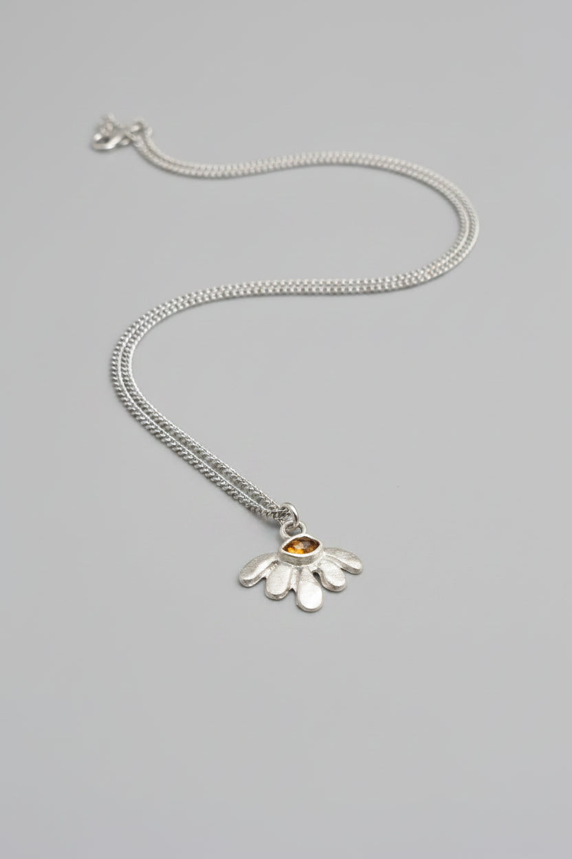 A sterling silver pendant necklace featuring a textured, petal-like daisy design with a small amber-orange gemstone in the center, resting on a dark brown textured leather background.