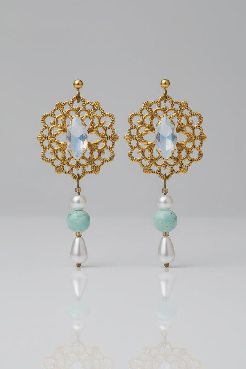 A pair of bohemian-style dangle earrings with ornate gold filigree, light blue marbled beads, and white pearl-like drops, displayed on a black card over a gray textured fabric background.