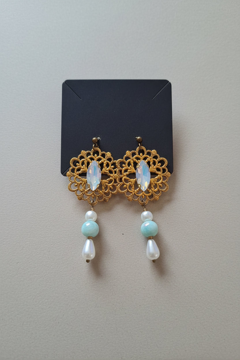 A pair of ornate gold filigree dangle earrings with iridescent oval stones, light blue beads, and white teardrop beads, displayed on a black card over a gray textured fabric background.