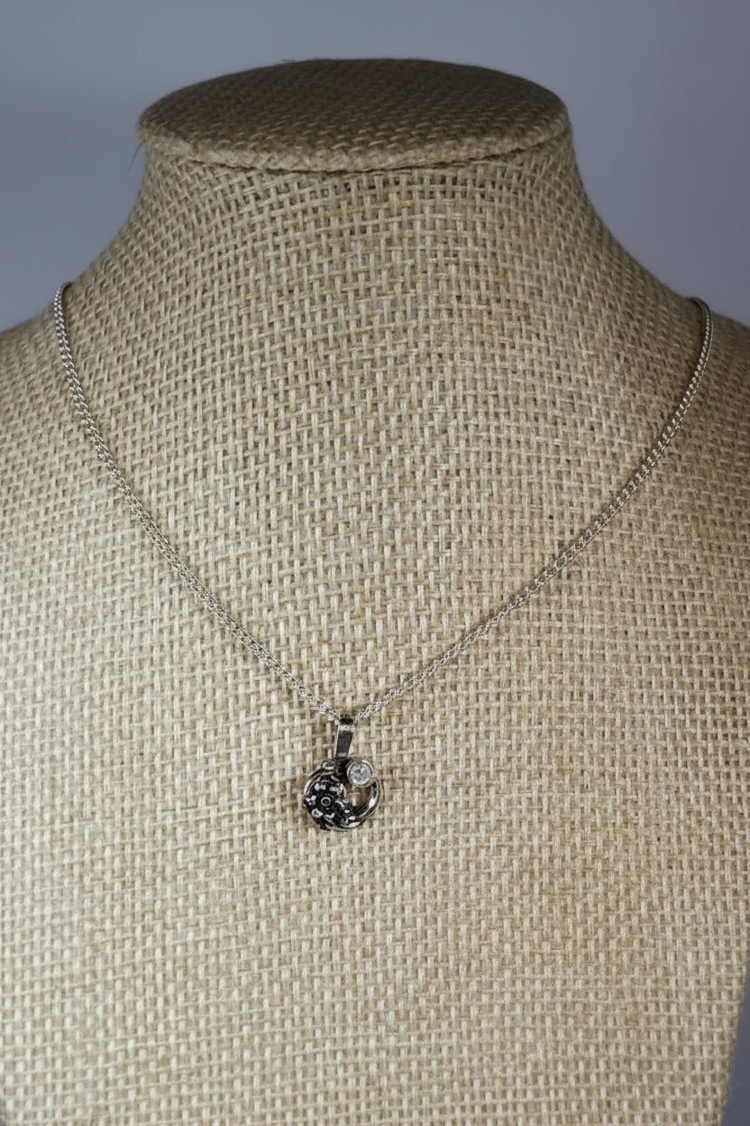 A silver-toned necklace with a small floral or snowflake-like pendant displayed on a woven burlap mannequin bust.