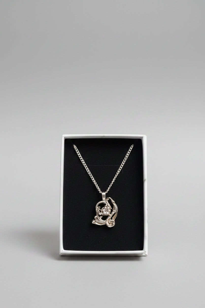 A silver-toned pendant necklace with a stylized animal or creature design, displayed inside an open white jewelry box with a black insert. A hand is holding the box.