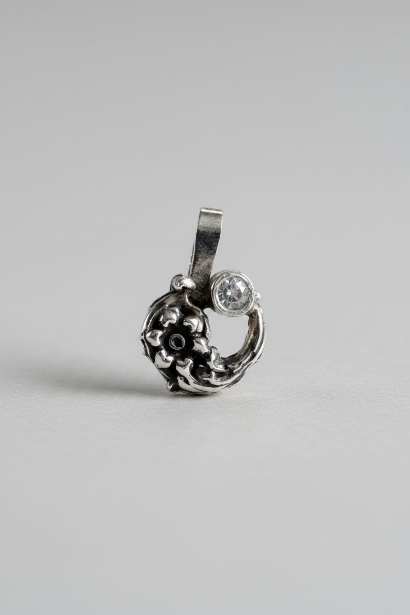 A small silver-toned pendant with an ornate floral or scroll design and a clear round stone, shown on a textured beige fabric background.