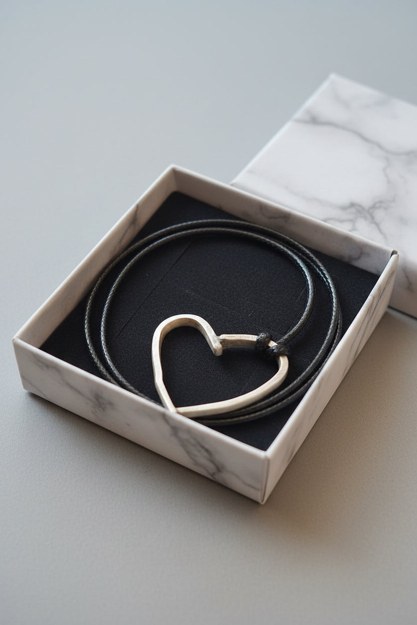 A silver-toned open heart pendant necklace with a black cord, displayed inside a black velvet-lined gift box.