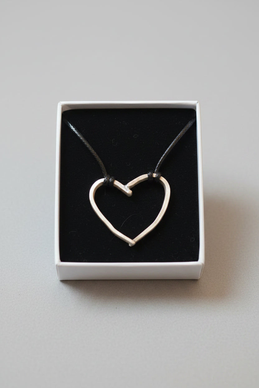 A silver heart-shaped pendant necklace on a black cord, presented inside an open white box with a black velvet insert.