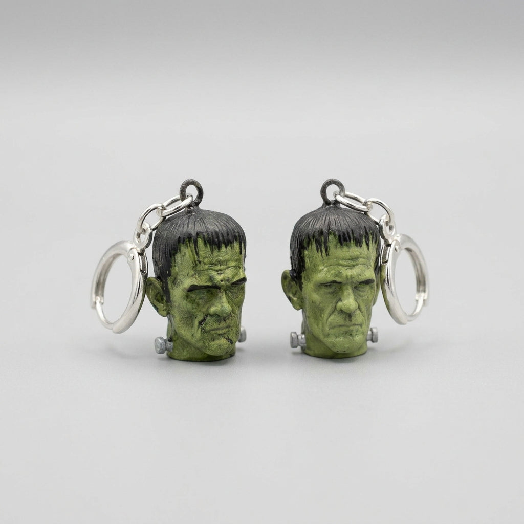 A pair of dangling earrings shaped like Frankenstein's monster heads, featuring green skin and black hair, with silver-tone rings, displayed on a textured gray fabric background.