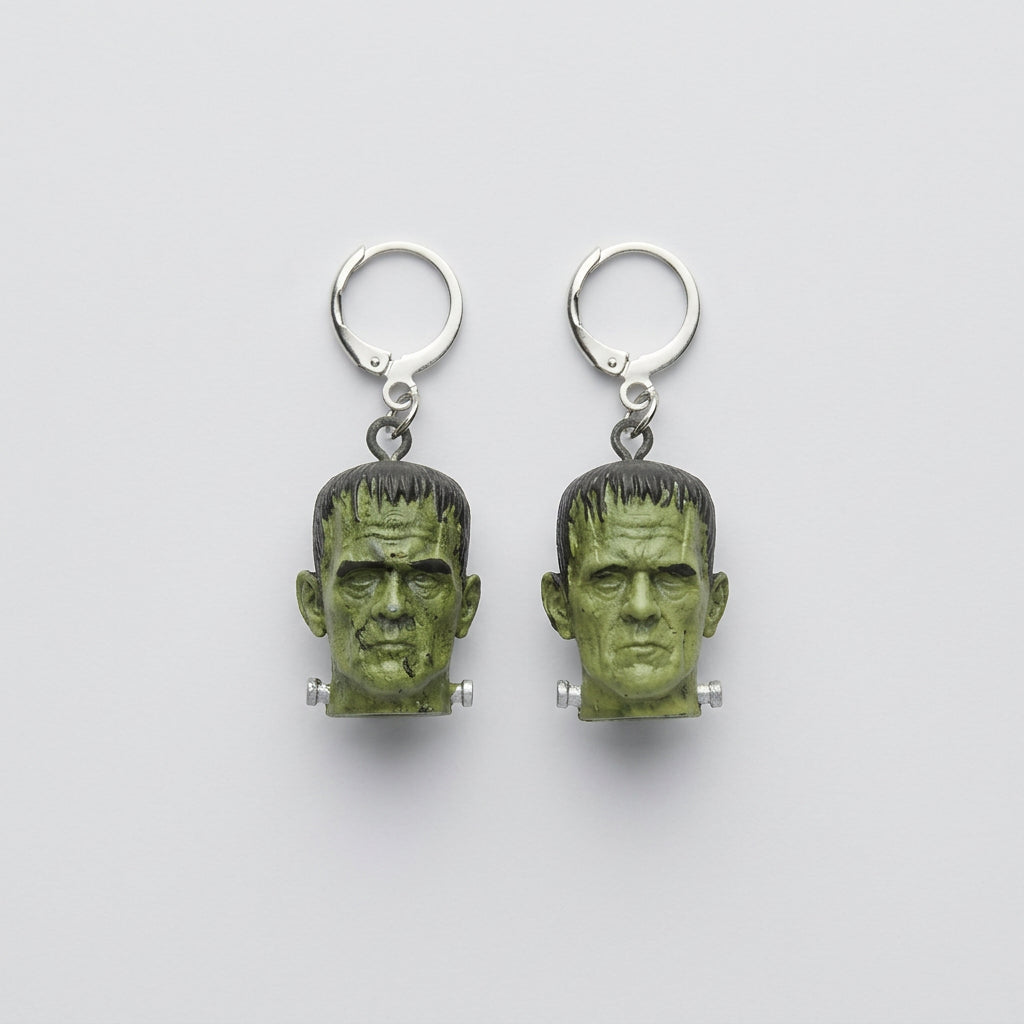 A pair of silver hoop earrings with dangling Frankenstein's green monster head charms, displayed on a textured gray fabric surface.