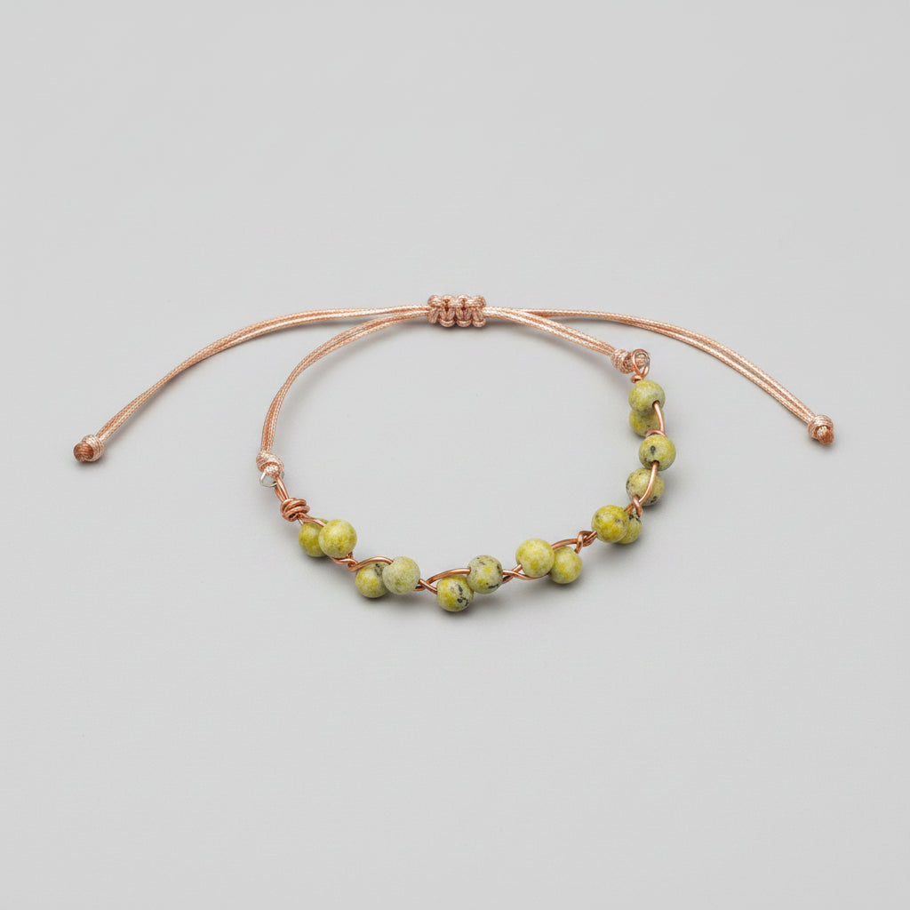 A handmade adjustable cord bracelet with small green and yellow marbled beads wrapped in copper wire, displayed on a textured gray fabric surface.