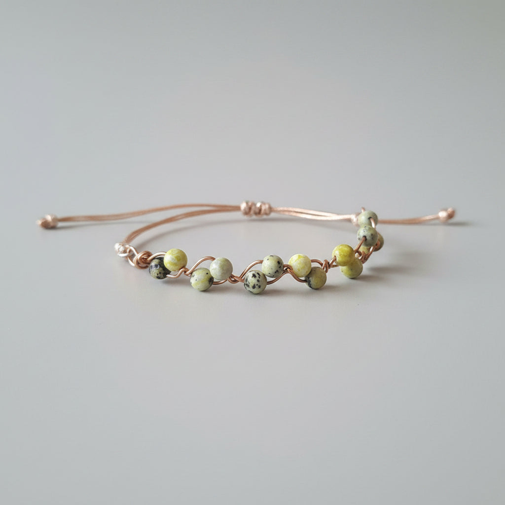 A handmade wire-wrapped bracelet with round beads in yellow-green and dark gray, resting on a textured gray fabric surface.