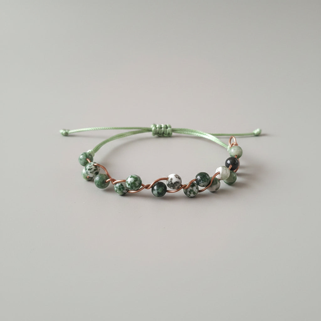 A handmade beaded bracelet with green and white marbled beads connected by copper wire, resting on a textured gray fabric surface.