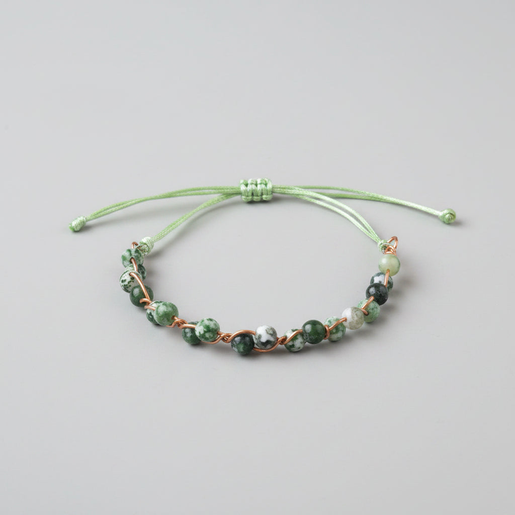 A handmade beaded bracelet with green cord, green and white marbled beads, and copper wire accents, resting on a gray textured cushion.