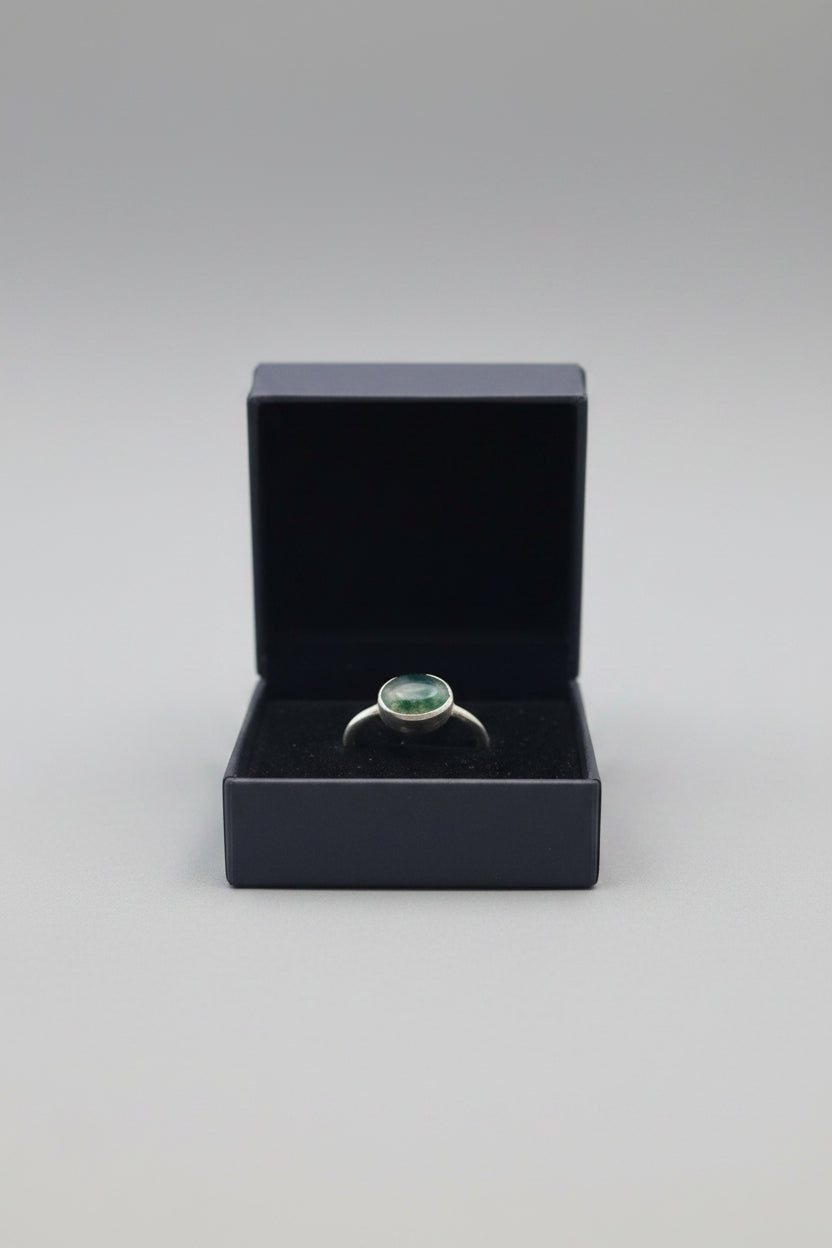 A silver ring with an oval green stone, presented in a black velvet box held by a hand.