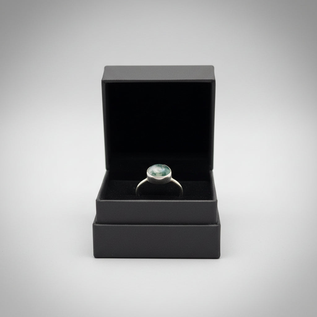 A silver ring with a round, moss agate stone displayed in a black velvet ring box, held by a hand in a home setting.