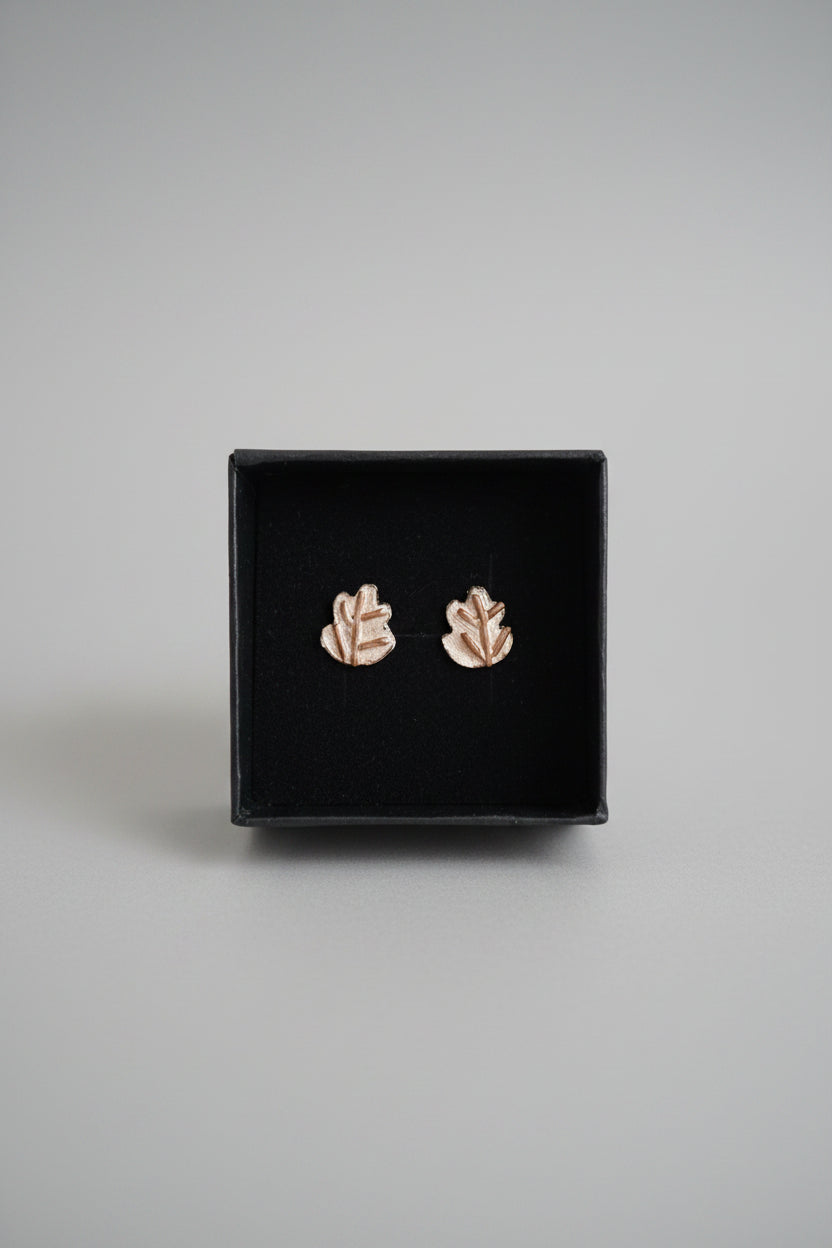 A pair of silver and copper leaf stud earrings displayed on a black presentation box, held by a hand in front of a blurred laptop keyboard and screen.