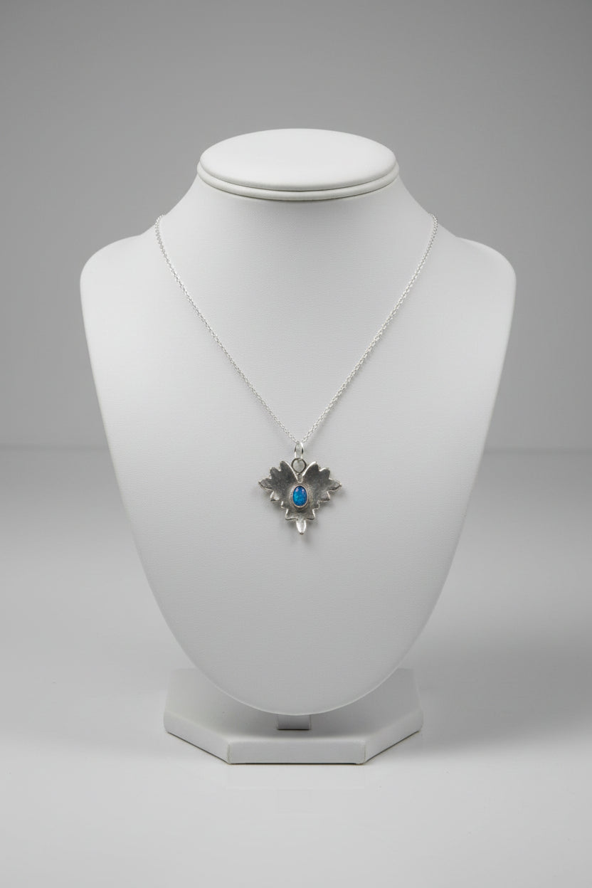 A silver-toned pendant necklace with a textured, leaf-like silhouette and a small dark blue oval stone in the center, displayed on a tan burlap-covered mannequin bust.