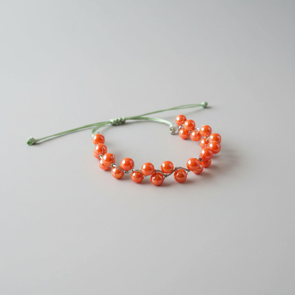 A beaded bracelet with glossy orange-red translucent beads on a thin green cord, resting on a textured gray fabric surface.