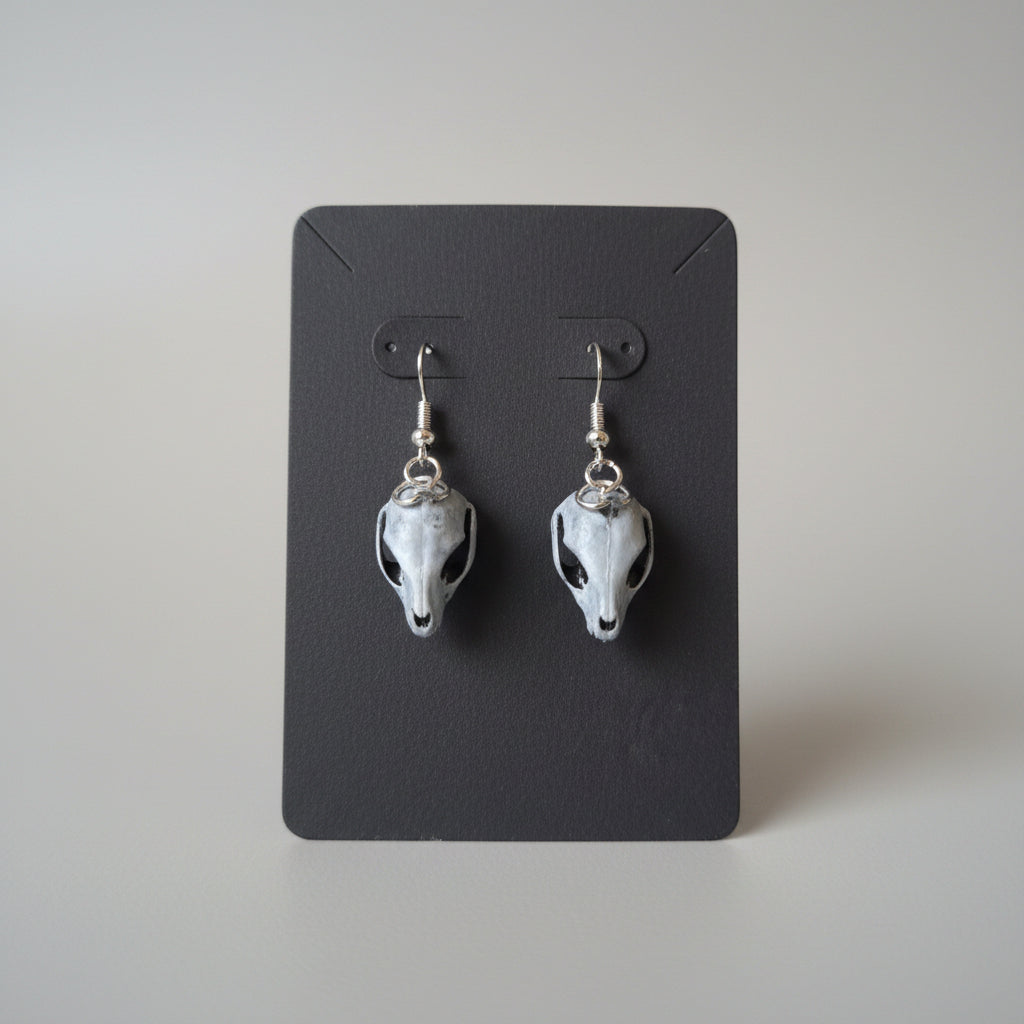 A pair of animal skull-shaped dangle earrings with silver-toned hooks, resting on a black card over a gray textured fabric surface.