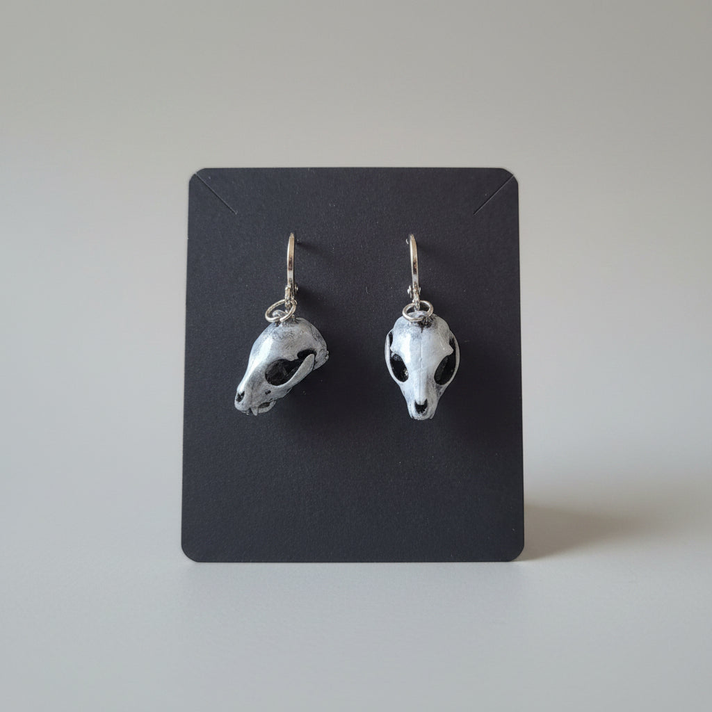 A pair of animal skull dangle earrings displayed on a black card, resting on a textured gray blanket in a home setting.