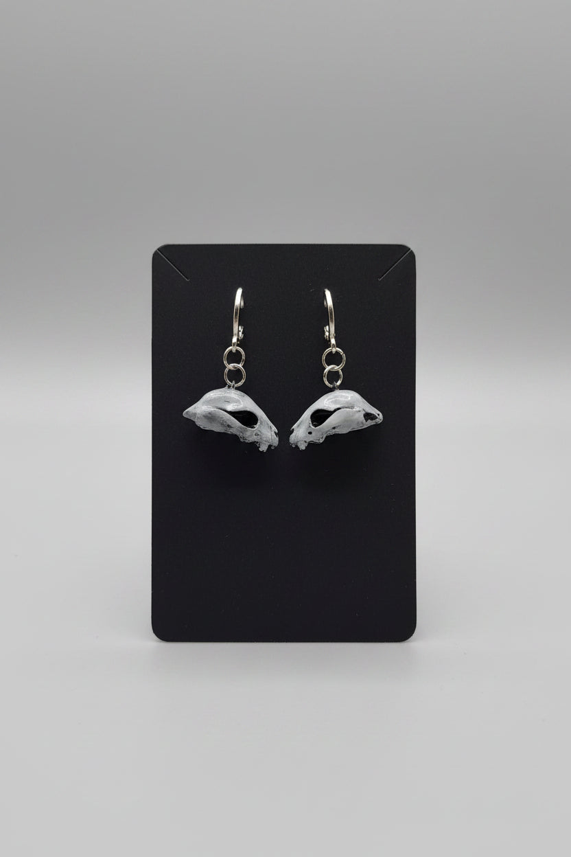 A pair of silver-toned drop earrings shaped like animal skulls, displayed on a black card held by a hand. The background is a blurry indoor setting.