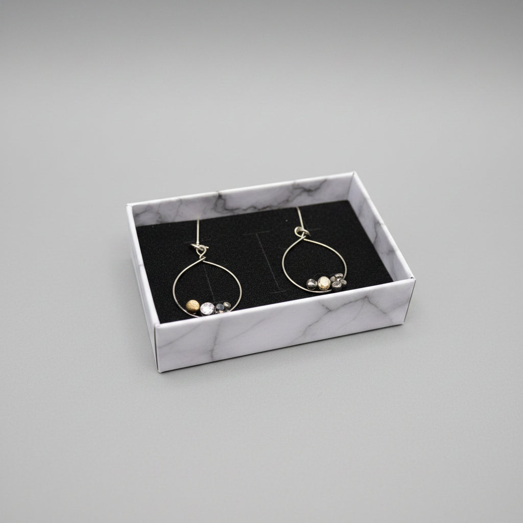 A pair of silver-tone hoop dangle earrings with small clear faceted beads, presented in an open white jewelry box with a black velvet insert, set on a gray textured fabric surface.