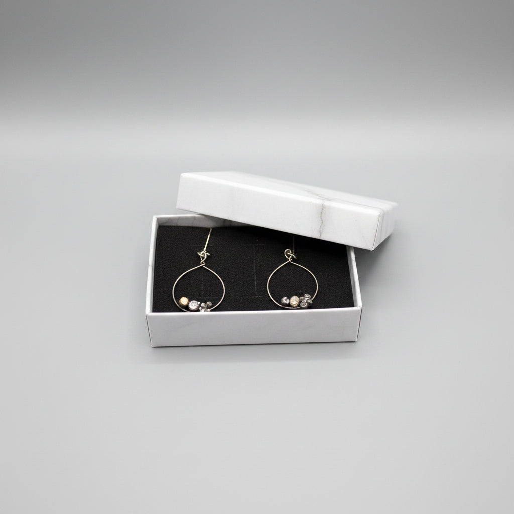 A pair of silver-tone hoop dangle earrings with small clear faceted beads, displayed inside an open white jewelry box with a black velvet lining, set on a gray textured fabric surface.