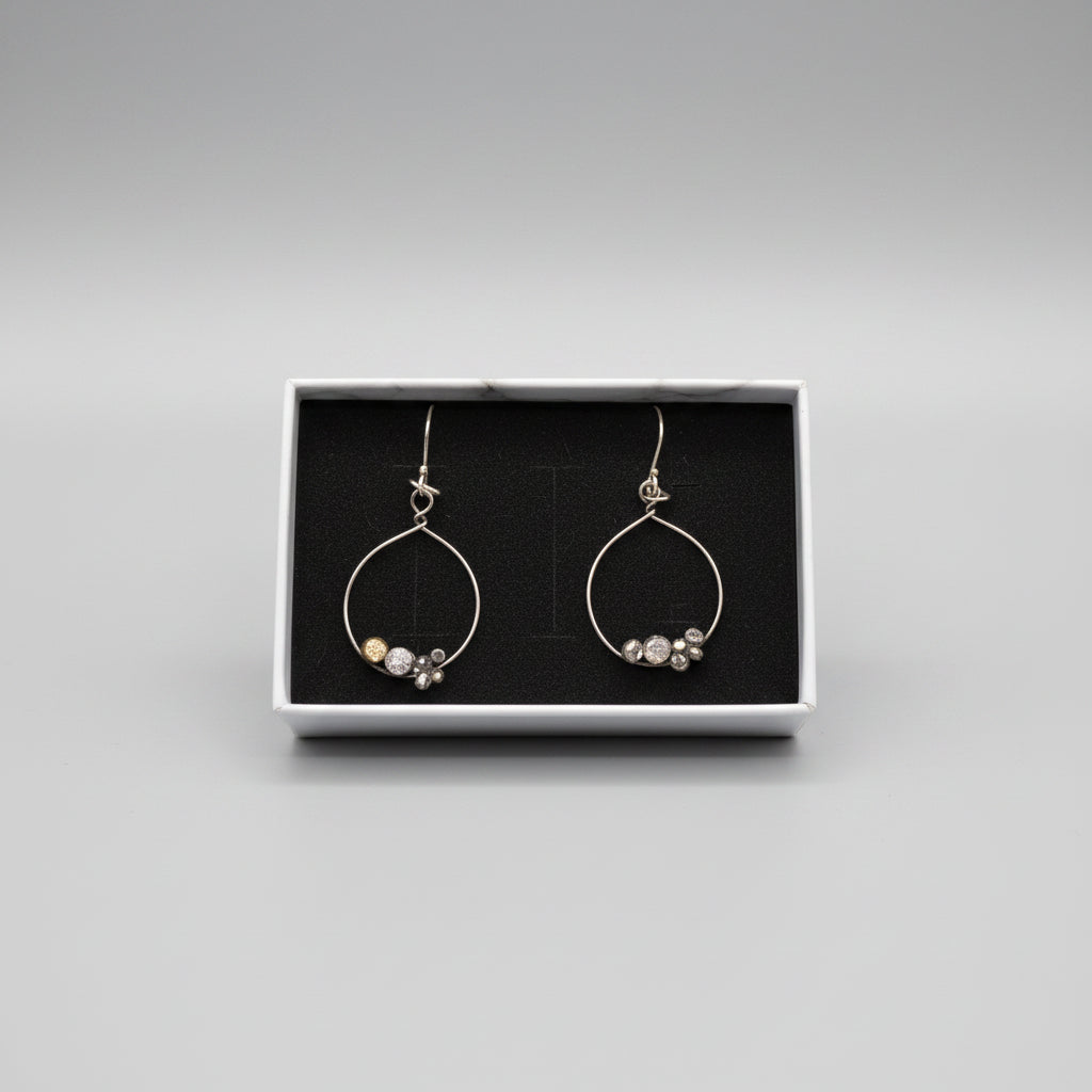 A pair of silver hoop dangle earrings with small clear and metallic beads, presented in a white box with a black insert, placed on a textured gray fabric surface.