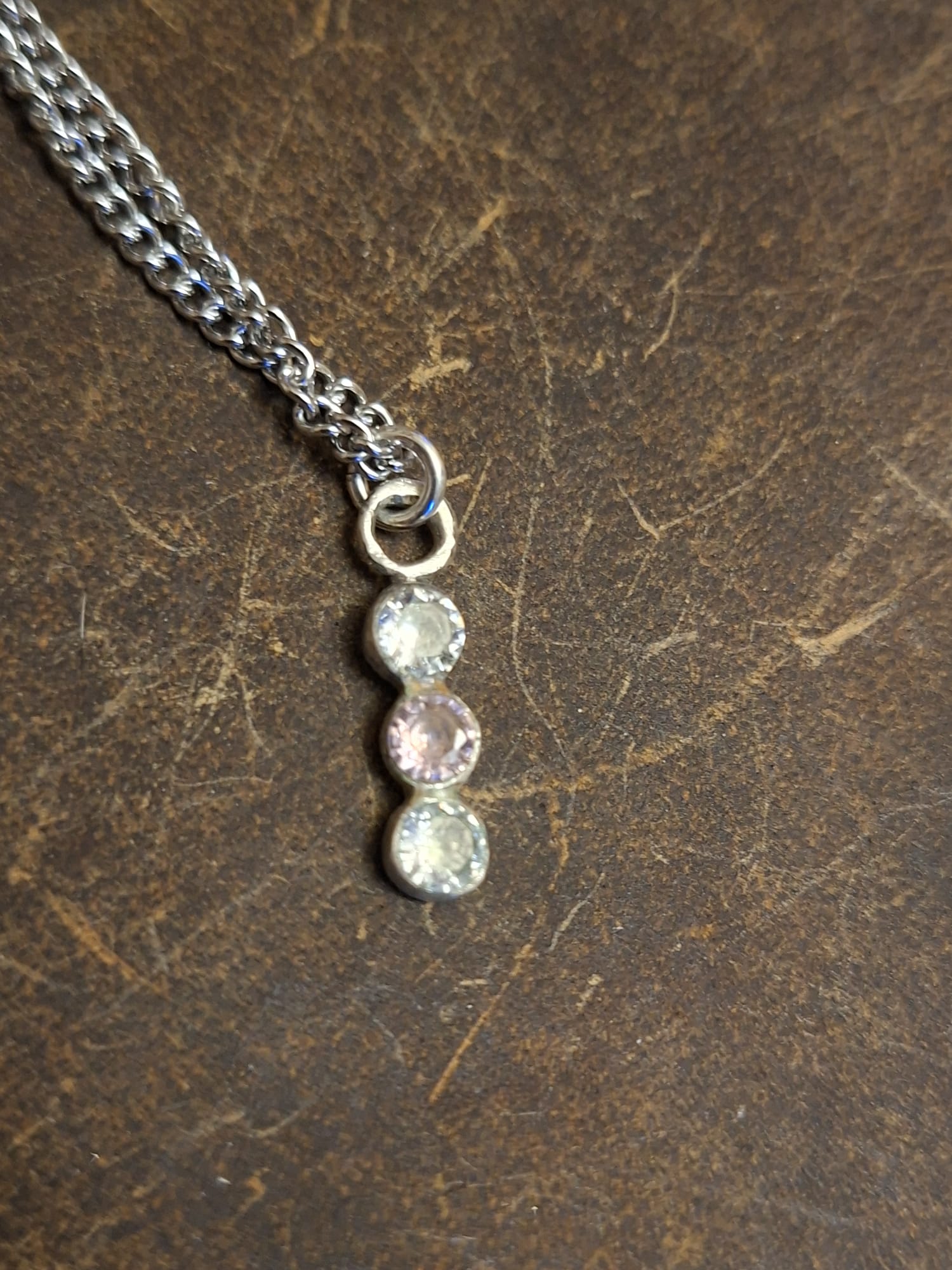 A close-up of a silver chain necklace with a vertical dangle charm featuring three round gemstones in silver settings, resting on a textured dark brown surface.