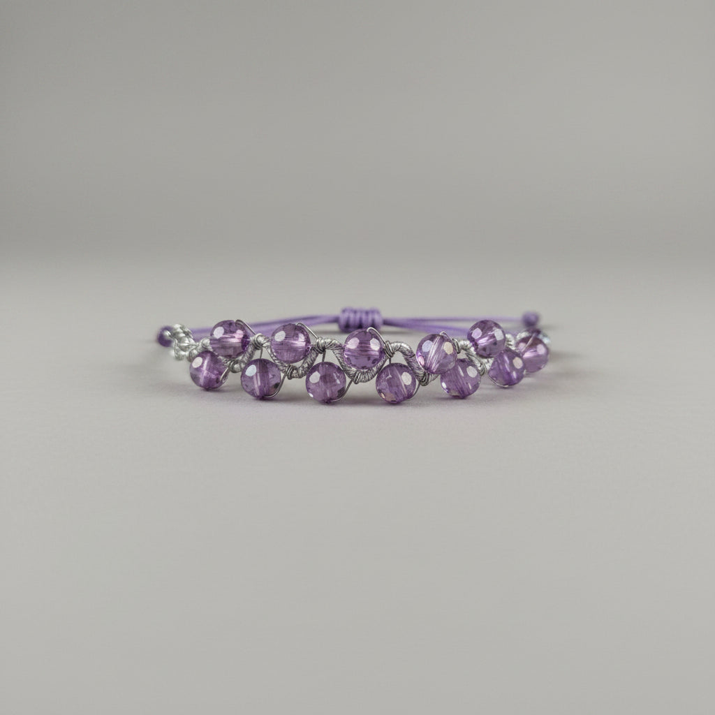 A handmade beaded bracelet with faceted light purple beads on a cord, resting on a textured gray fabric surface.